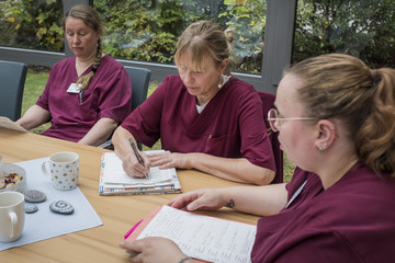 Drei Fachpflegerinnen in weinroter Funktionskleidung sitzen auf der Terrasse an einem Tisch mit Dokumenten bei einer Besprechung im Diakonie Hospiz Volksdorf in Hamburg-Volksdorf. 