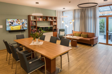 Blick in den wohnlich eingerichteten hellen Aufenthaltsraum mit Holzfußboden, indirekter Beleuchtung, einem großen Tisch mit Stühlen, Sofa, Regal und großem Monitor im Diakonie Hospiz Volksdorf in Hamburg-Volksdorf.