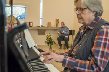Eine ehrenamtliche Helferin spielt an einem Klavier in Begleitung von Seelsorger Roger Bahr in einem festlich geschmückten Raum mit einem Fenster in Kreuzform im Diakonie Hospiz Volksdorf in Hamburg-Volksdorf. 