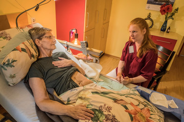 Eine Fachpflegerin in weinroter Funktionskleidung sitzt neben dem Pflegebett im Gästezimmer einer Hospizgästin und unterhält sich mit ihr im Diakonie Hospiz Volksdorf in Hamburg-Volksdorf.
