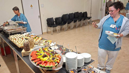 Zwei Pflegerinnen in blauer Funktionskleidung stehen in einem Raum mit einem mit warmen und kalten Speisen, Obst, Müsli und Gebäck gedeckten Buffet am Internationalen Tag der Pflegenden.