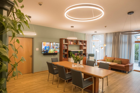 Blick in einen Gemeinschaftsraum mit Holzboden, einem mit Blumen geschmückten Tisch, Stühlen, Sofa und Regal, einem großen Monitor und modernen Deckenlampen im Diakonie Hospiz Volksdorf in Hamburg-Volksdorf.