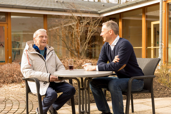 Zwei Menschen sitzen am Terrassentisch. Der Hospizgast unterhält sich mit einem Mitarbeiter. Zwei Menschen sitzen am Terrassentisch. Der Hospizgast unterhält sich mit einem Mitarbeiter.